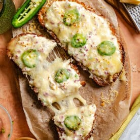 Three slices of toasted bread topped with melted cheese, sliced jalapeños, and bits of ham, served on parchment paper with a halved jalapeño and a block of cheese nearby.