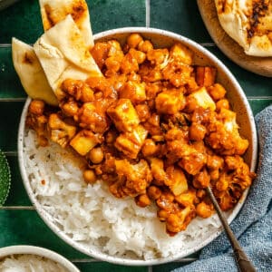 A bowl of white rice topped with chickpea and paneer curry, garnished with naan bread, sits on a green tiled surface with a spoon resting in the bowl.