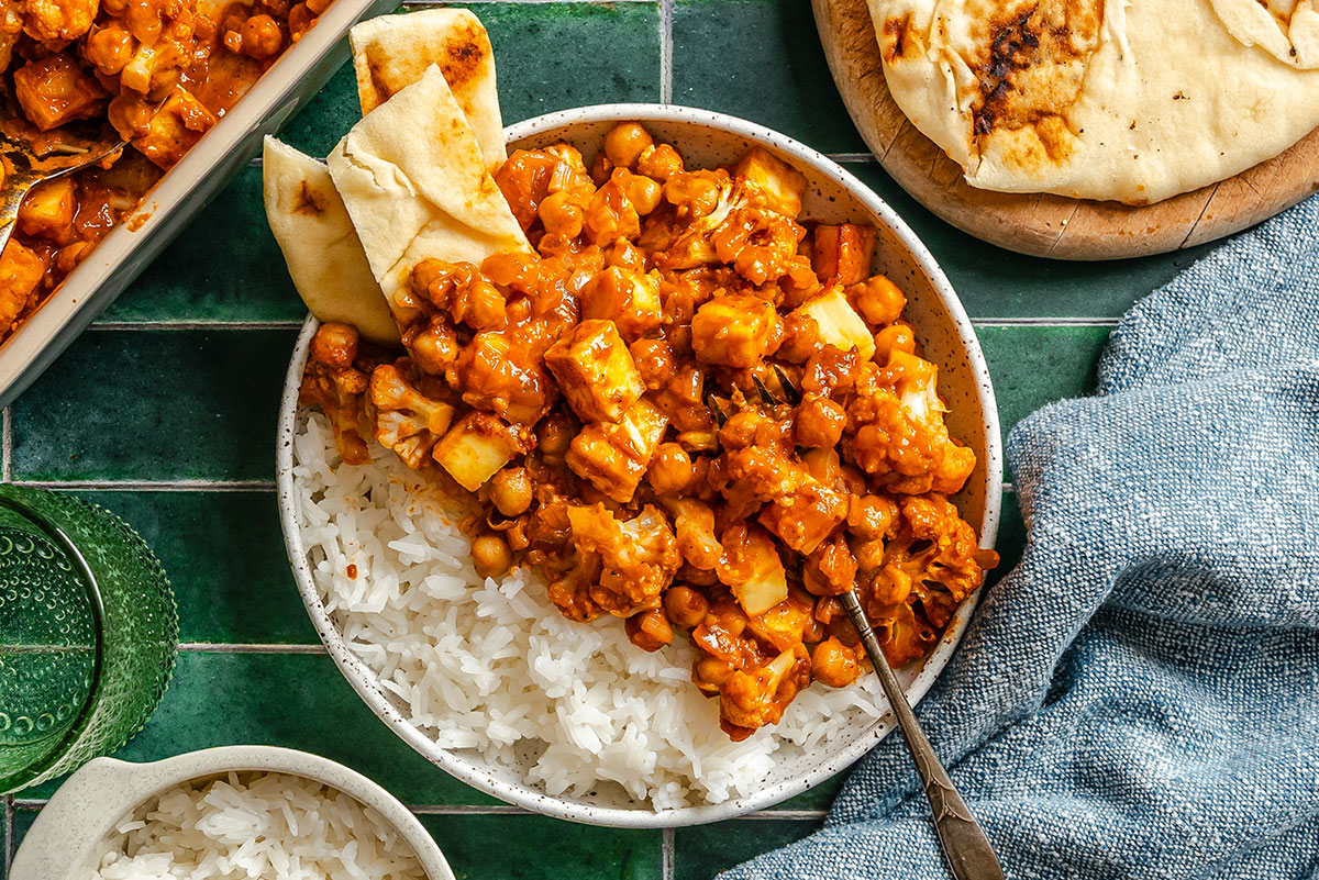 A bowl of white rice topped with a chickpea and paneer curry, garnished with naan bread, on a green tiled surface with a blue cloth napkin.