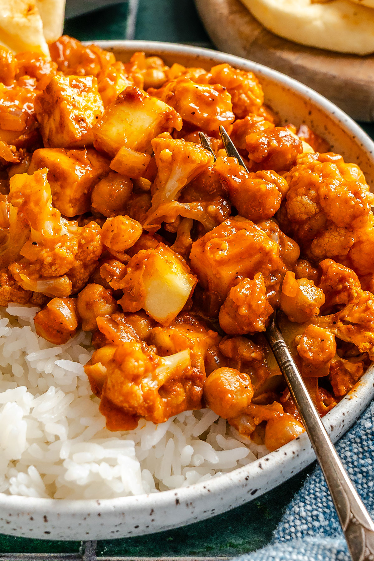 A bowl of white rice topped with a chickpea, cauliflower, and potato curry in a rich orange sauce, with a spoon resting in the bowl.