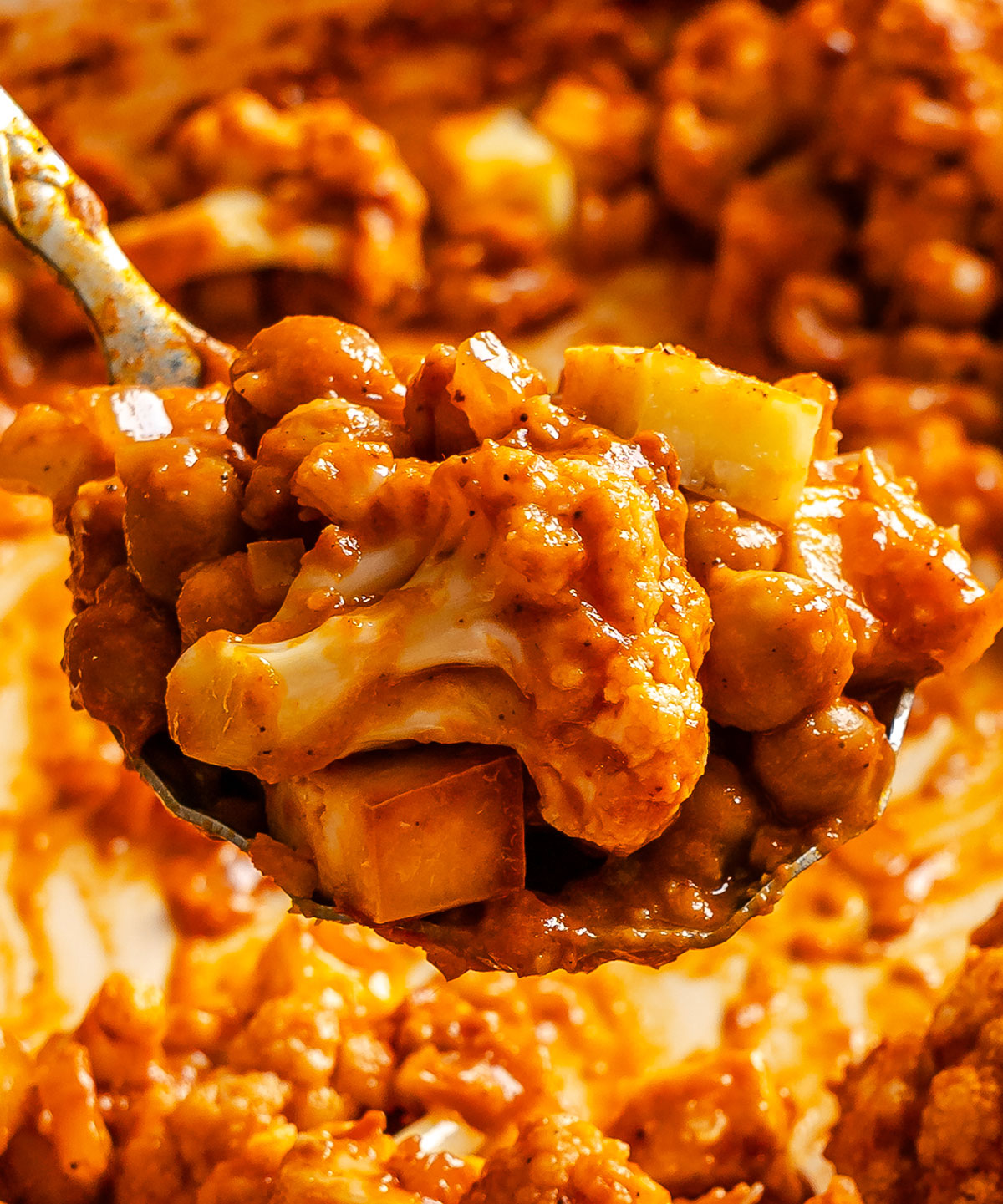 A close-up of a spoonful of curry featuring cauliflower, chickpeas, and potato cubes in a thick, orange sauce.