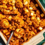 Baked dish with cauliflower florets, chickpeas, cubed paneer, and sauce, served in a rectangular baking dish with a metal serving spoon.