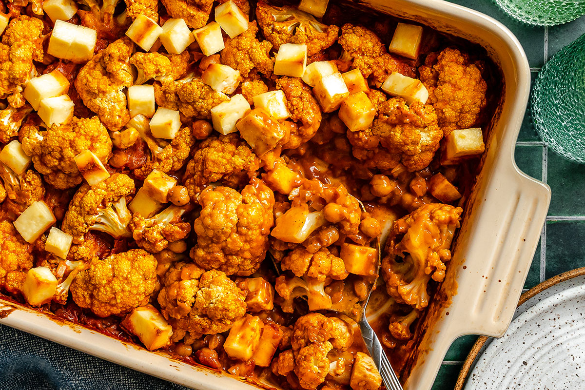 Baked cauliflower florets and chickpeas in a reddish sauce, topped with diced cheese, in a rectangular baking dish with a serving fork.