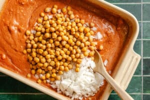 A baking dish with a tomato-based sauce, topped with chickpeas and chopped onions, being mixed with a wooden spoon on a green tiled surface.