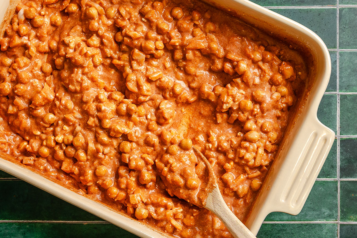 A baking dish filled with chickpea curry in a creamy tomato sauce, with a wooden spoon resting inside the mixture.
