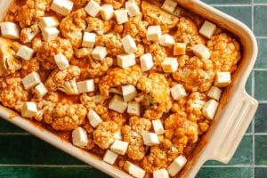A baking dish filled with cauliflower florets, chickpeas, tomato sauce, and topped with cubed paneer cheese on a green tiled surface.