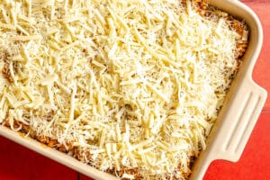 A rectangular baking dish filled with an unbaked casserole topped with a generous layer of shredded cheese on a red tiled surface.