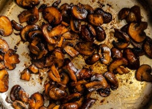 Sliced mushrooms sautéed in a stainless steel pan, showing browned edges and caramelization.