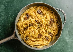A skillet filled with cooked spaghetti and sautéed mushrooms on a green surface.