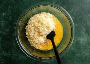 A glass bowl containing beaten eggs and grated cheese, with a black spatula resting inside, on a green countertop.