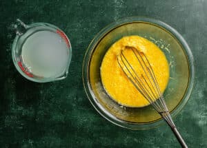 A glass bowl with a yellow egg mixture and a whisk next to a measuring cup filled with a cloudy liquid on a green countertop.