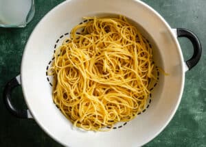 Cooked spaghetti noodles draining in a white colander with black handles, placed on a green countertop.