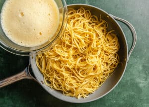 A glass bowl of beaten eggs is positioned above a pan filled with cooked spaghetti on a green surface.