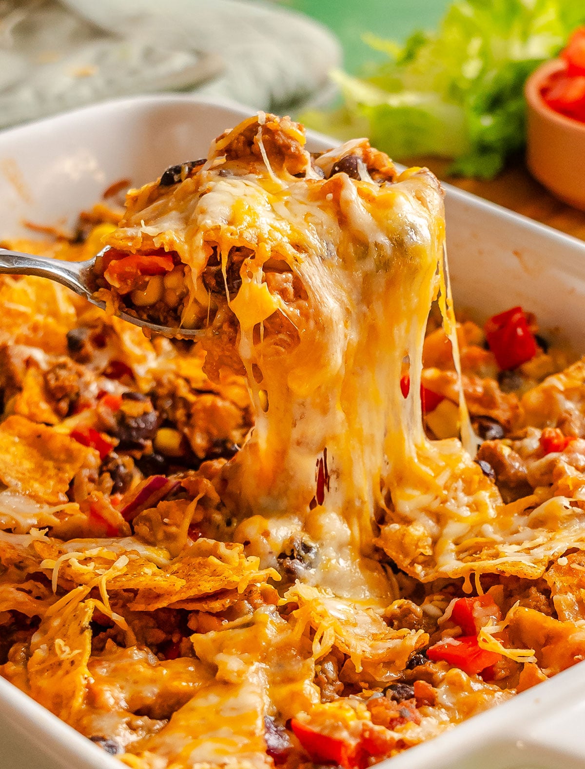 A cheesy nacho casserole with melted cheese, beans, corn, and chopped red peppers being served from a white baking dish.