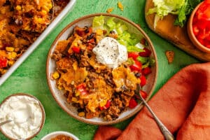 A bowl of taco casserole topped with sour cream, served with lettuce and diced tomatoes on a green table with an orange napkin and extra sour cream on the side.