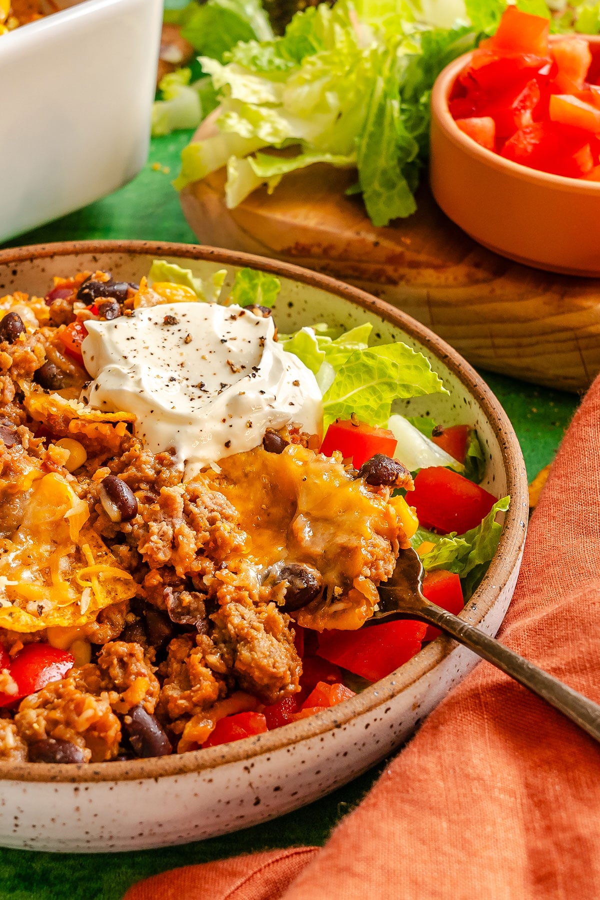 A bowl of taco casserole with ground meat, black beans, melted cheese, chopped lettuce, tomatoes, and a dollop of sour cream, with a fork resting inside.