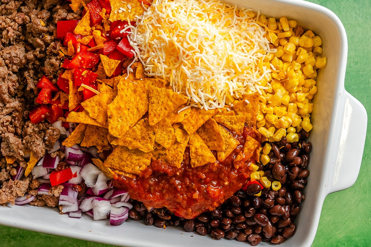 A casserole dish filled with ground beef, chopped red bell pepper, shredded cheese, corn, black beans, red onion, salsa, and tortilla chips, arranged in separate sections.