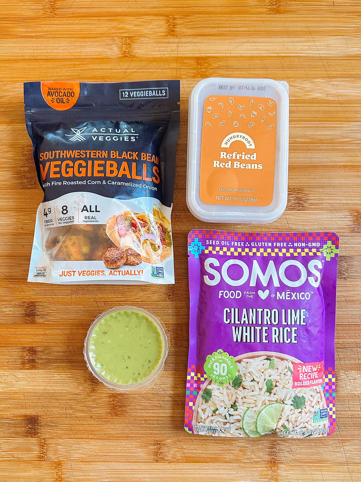 Packaged southwestern black bean veggieballs, a container of refried red beans, cilantro lime white rice, and a small cup of green sauce arranged on a wooden surface.
