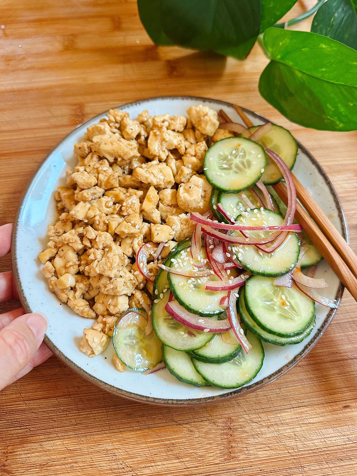 A plate with crumbled tofu and a cucumber salad with red onions and sesame seeds, placed on a wooden surface with a pair of chopsticks and green leaves in the background.