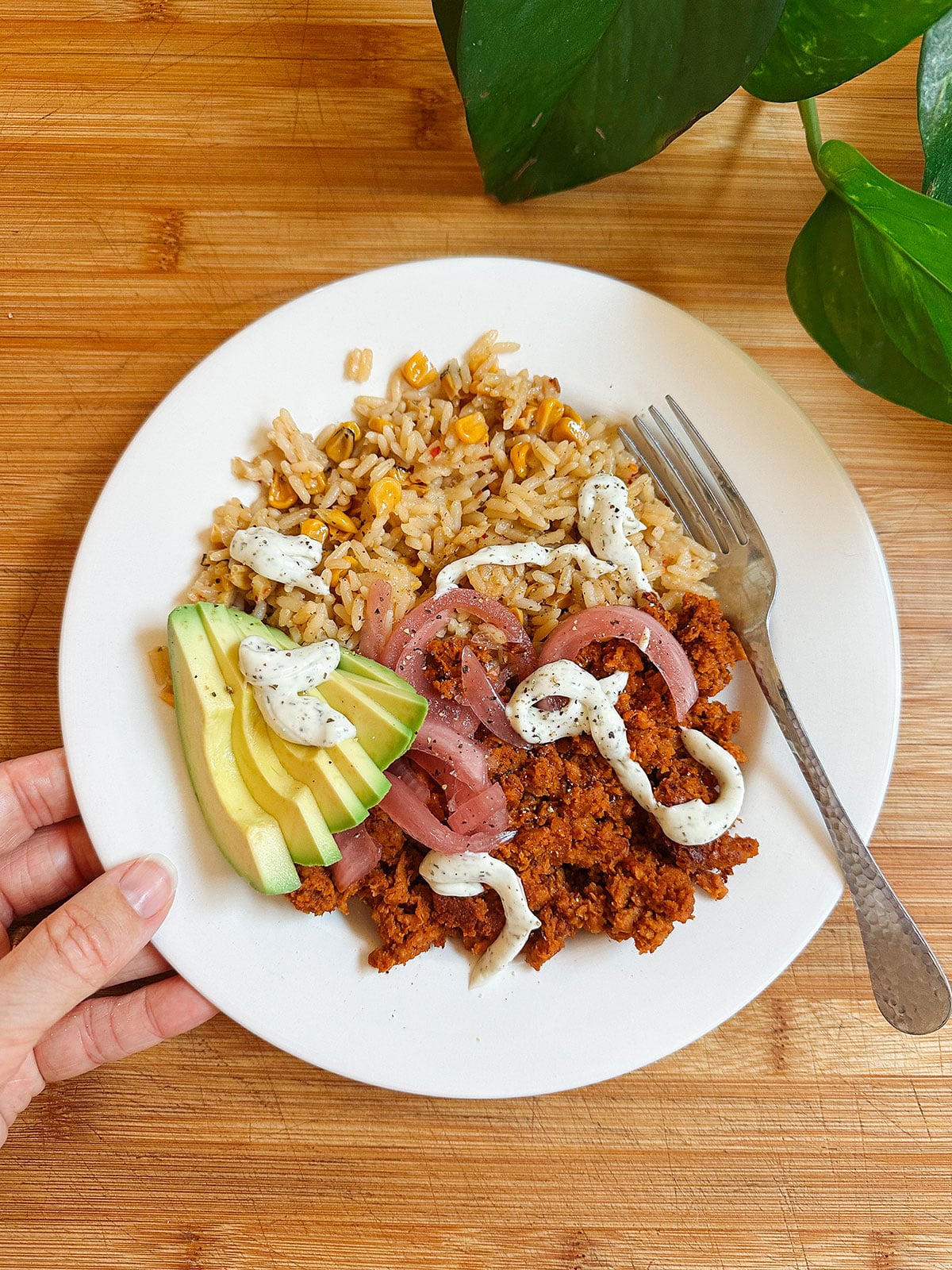A plate with seasoned rice and corn, sliced avocado, pickled onions, plant-based ground meat, and a drizzle of creamy sauce. A fork rests on the plate beside the food.