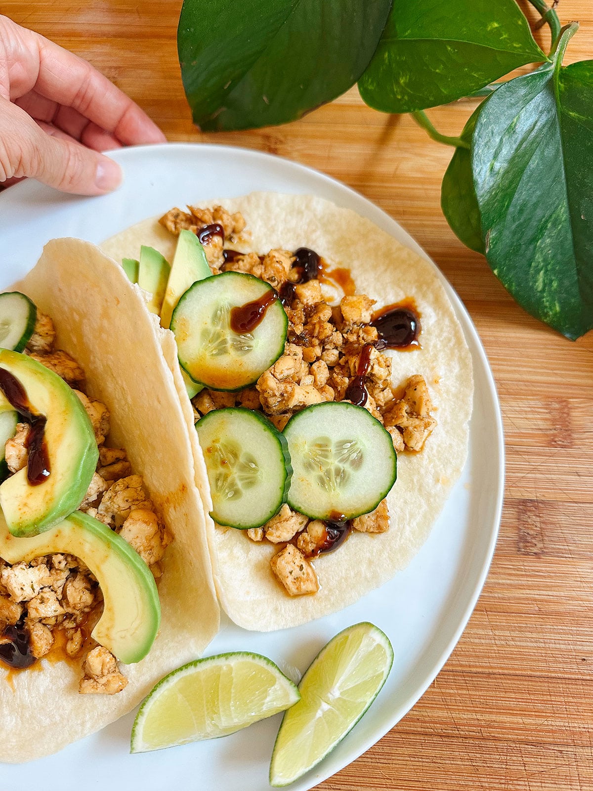 Two soft tacos on a white plate filled with diced tofu, cucumber slices, avocado, and sauce, served with lime wedges; a hand touches the plate, and a green plant is nearby.