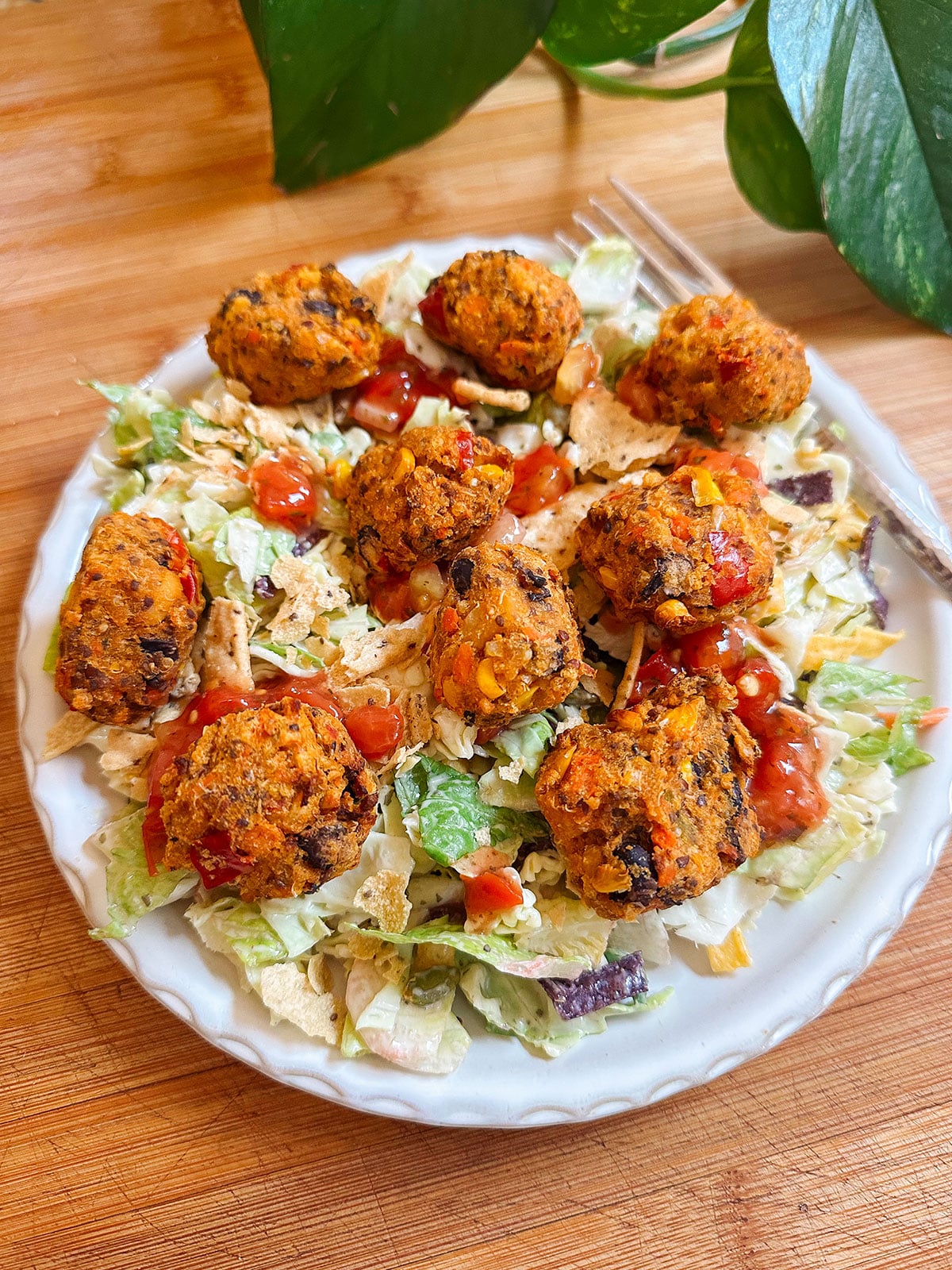 A plate of salad topped with several baked veggie balls and drizzled with red sauce, set on a wooden surface with a green plant in the background.