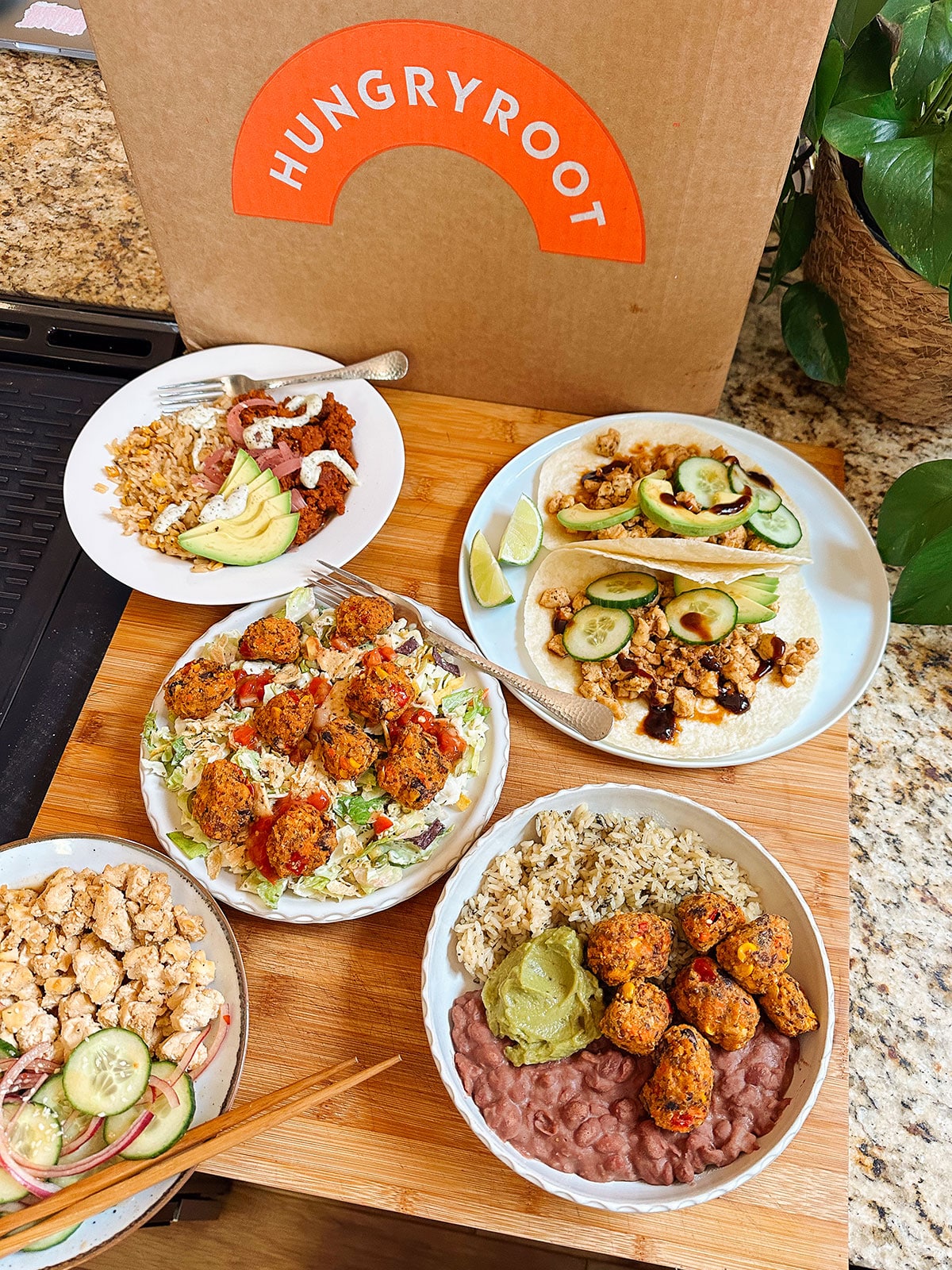 Five plated meals with vegetables, grains, and plant-based proteins are arranged on a wooden board in front of a Hungryroot delivery box on a kitchen counter.