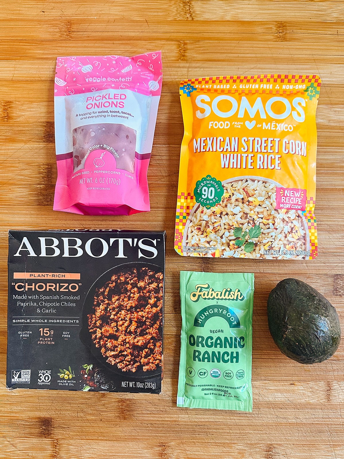 Five food items on a wooden surface: pickled onions, Mexican street corn white rice, plant-based chorizo, organic ranch dressing, and an avocado.