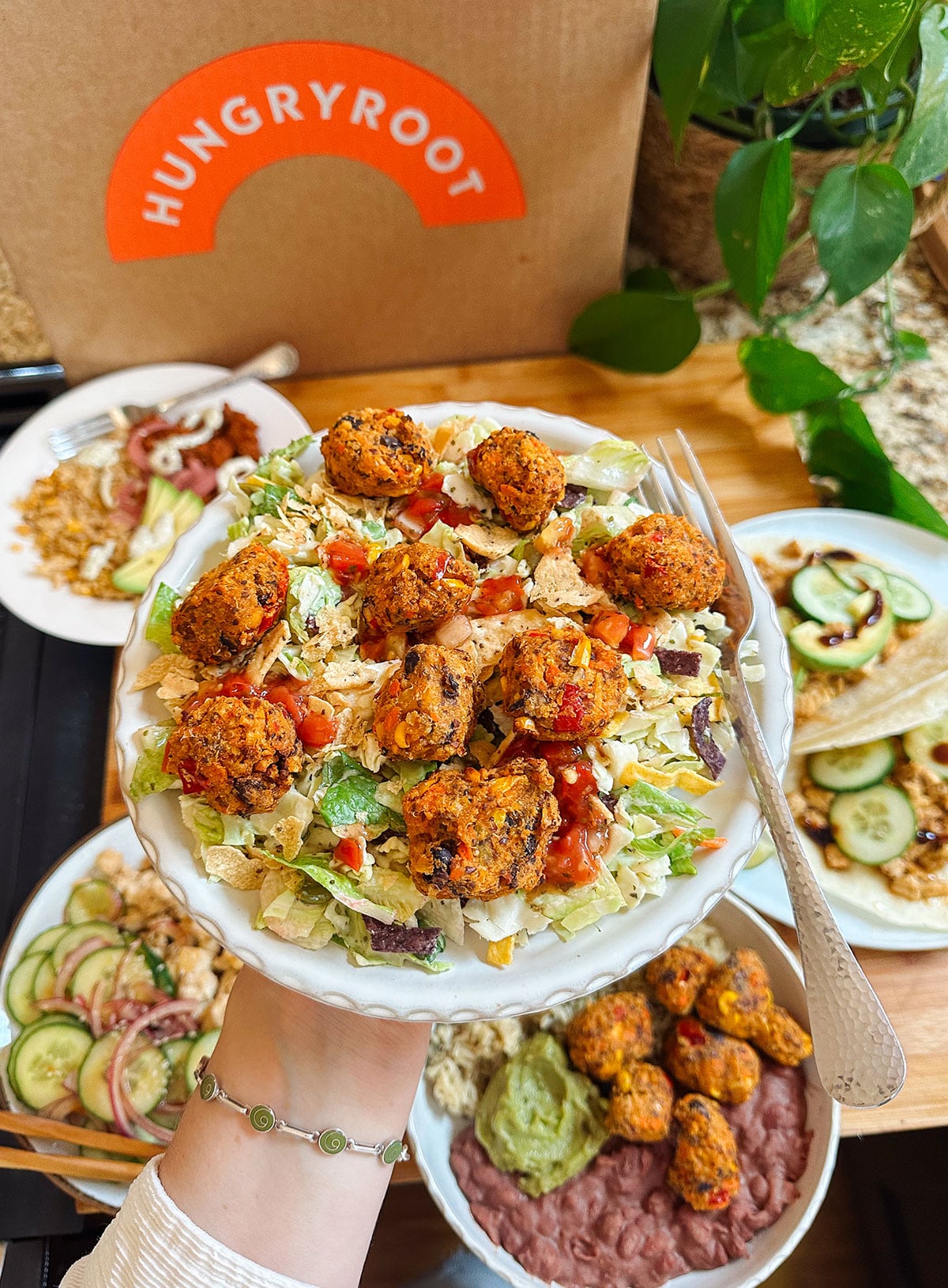 A hand holds a bowl of salad topped with falafel and sauce, with plates of various fresh dishes and a Hungryroot box in the background.