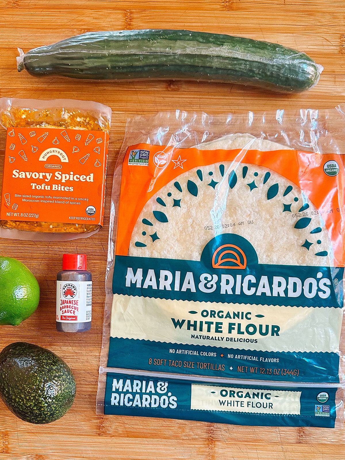 A cucumber, savory spiced tofu bites, a lime, Japanese chili powder, an avocado, and a package of organic white flour tortillas on a wooden surface.