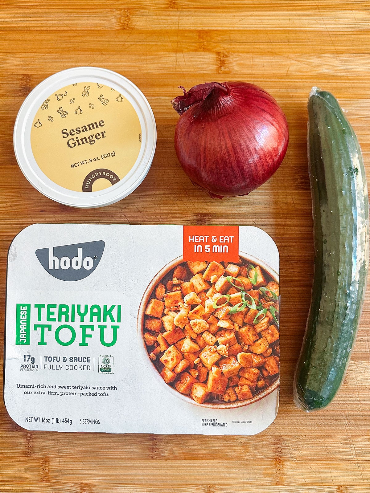 A container of sesame ginger sauce, a red onion, a cucumber, and a package of teriyaki tofu are arranged on a wooden cutting board.
