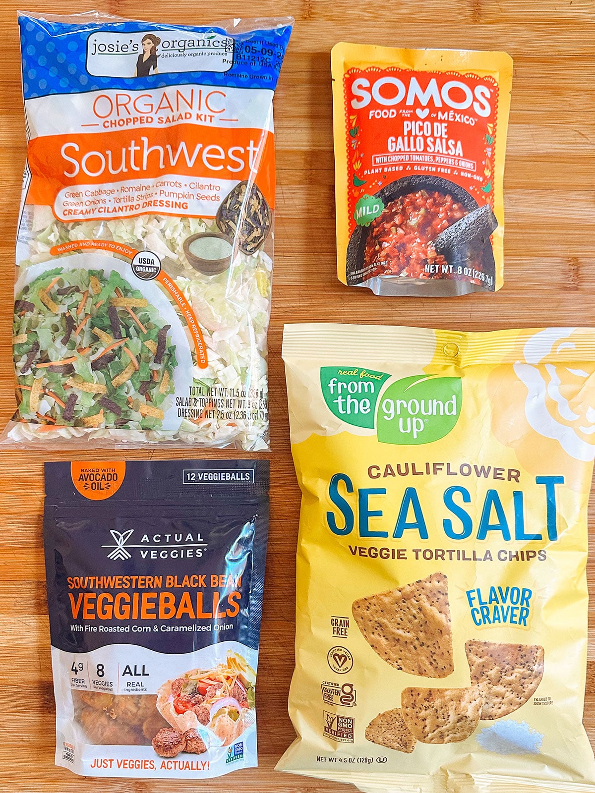 Four packaged food items on a wooden surface: a Southwest salad kit, pico de gallo black beans, black bean veggie balls, and cauliflower sea salt tortilla chips.
