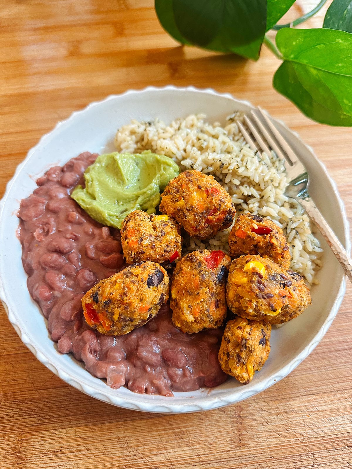 A bowl with refried beans, brown rice, guacamole, and several vegetable fritters, placed on a wooden surface.