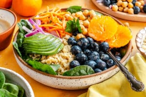 A bowl filled with quinoa, spinach, avocado slices, shredded carrots, red onion, chickpeas, blueberries, and orange slices, with a fork resting on the side.