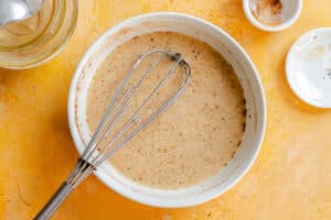 A metal whisk resting in a bowl of mixed batter on a yellow surface, with small bowls containing spices and a glass jar nearby.