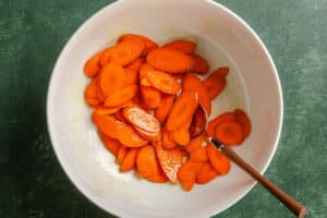 Sliced carrots coated in a light glaze, in a white bowl with a wooden-handled spoon on a green surface.