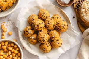 A plate of round, cookie dough balls with chocolate chips and a sprinkle of salt, surrounded by bowls of chickpeas, oats, and chocolate chips.
