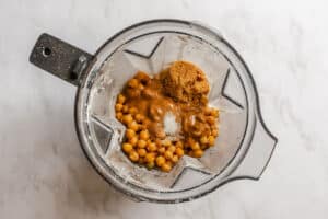 A blender containing chickpeas, brown sugar, nut butter, and salt, placed on a white surface.