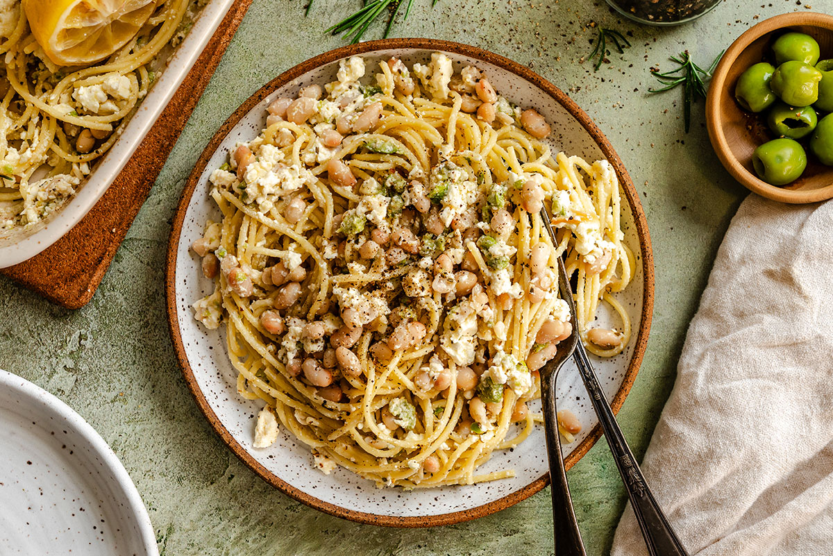 A plate of spaghetti topped with white beans and crumbled cheese, with black pepper, olives, and herbs visible nearby. Two forks are placed on the plate.