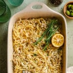 Baking dish filled with spaghetti mixed with white beans, sprinkled cheese, fresh rosemary, and a halved lemon. Green olives and glasses are nearby.