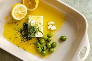 A baking dish with feta cheese, olive oil, green olives, garlic cloves, chopped herbs, and halved lemons arranged inside.