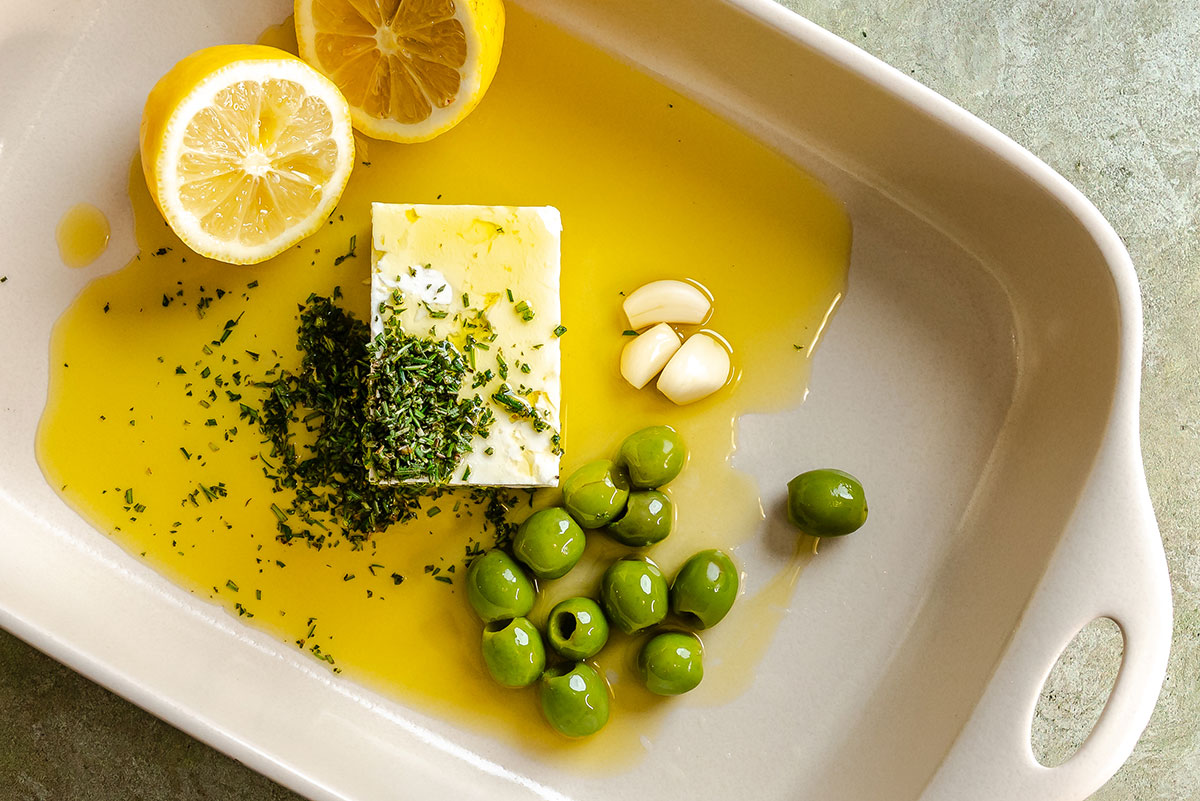 A baking dish with feta cheese, olive oil, green olives, garlic cloves, chopped herbs, and halved lemons arranged inside.