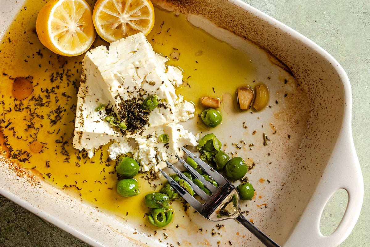 A baking dish with feta cheese, green olives, lemon halves, garlic cloves, herbs, and olive oil, with a fork resting in the dish.