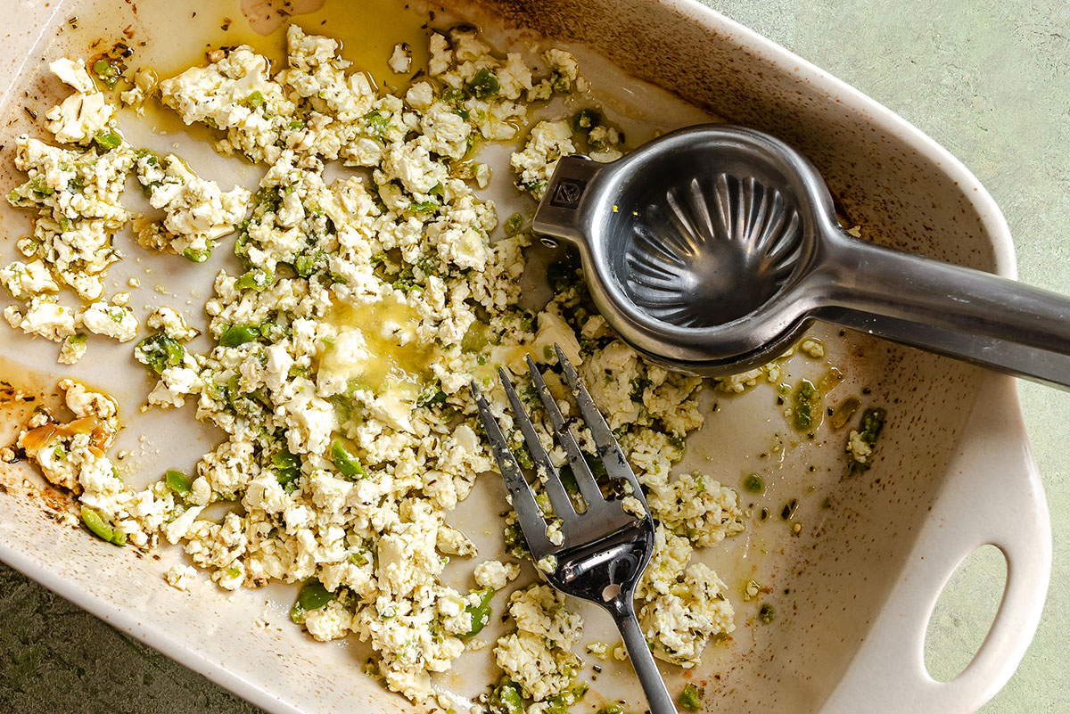 A baking dish with crumbled tofu or cheese mixed with herbs and oil, a metal citrus juicer, and a fork resting inside.