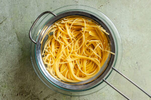 Cooked spaghetti draining in a metal strainer over a glass bowl on a gray textured surface.