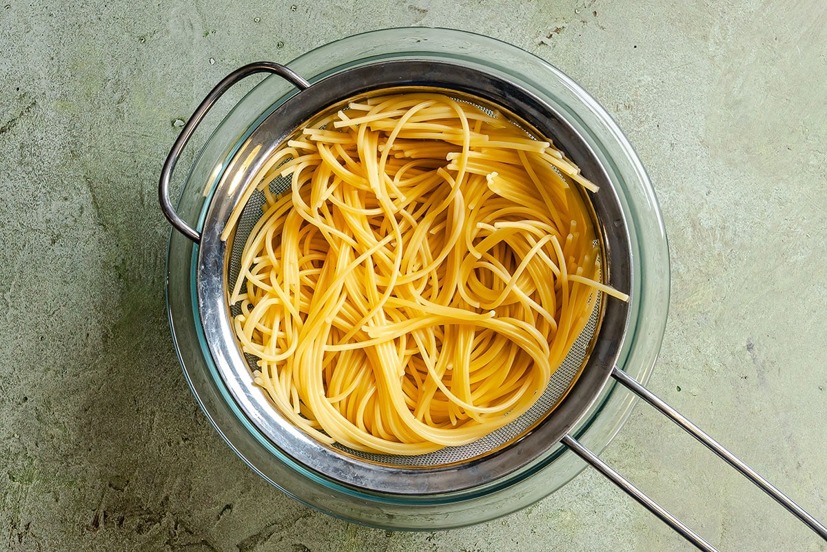 Cooked spaghetti draining in a metal strainer over a glass bowl on a gray textured surface.