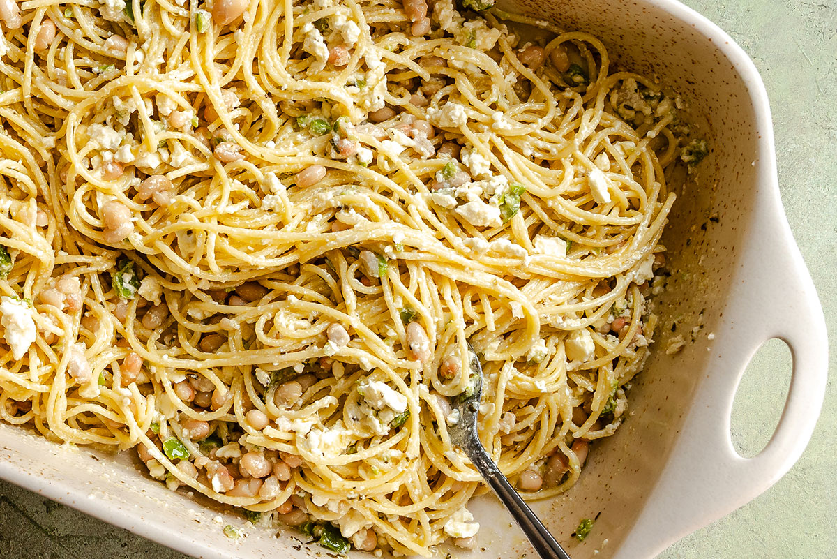 Baked spaghetti mixed with white beans, crumbled feta cheese, and herbs in a white ceramic dish with a serving fork.