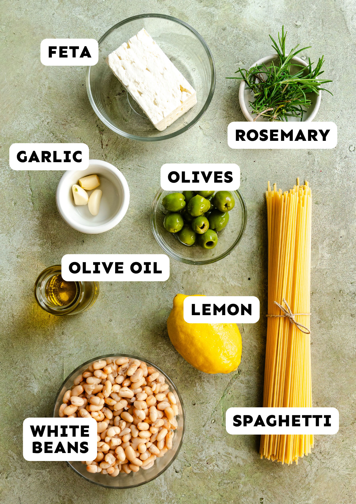 Top-down view of ingredients for Dirty Martini Spaghetti on a surface—feta, rosemary, garlic, olives, olive oil, lemon, spaghetti, and white beans—all labeled with text. Perfect for a Dump and Bake Martini Pasta night!.