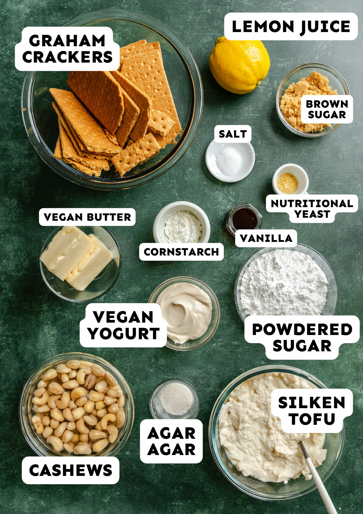 Various vegan cheesecake ingredients are arranged in bowls on a green surface, including graham crackers, silken tofu, cashews, vegan yogurt, powdered sugar, lemon, and labeled baking staples.