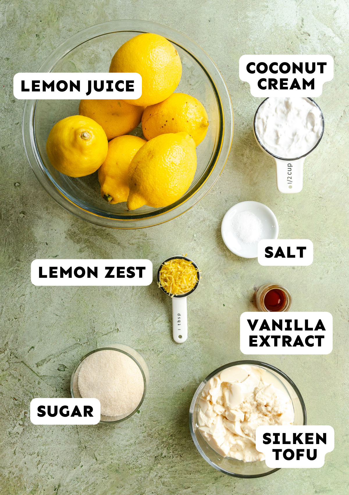 A top-down view of labeled ingredients on a green surface for Vegan Lemon Posset: lemons, coconut cream, salt, vanilla extract, lemon zest, sugar, and silken tofu.